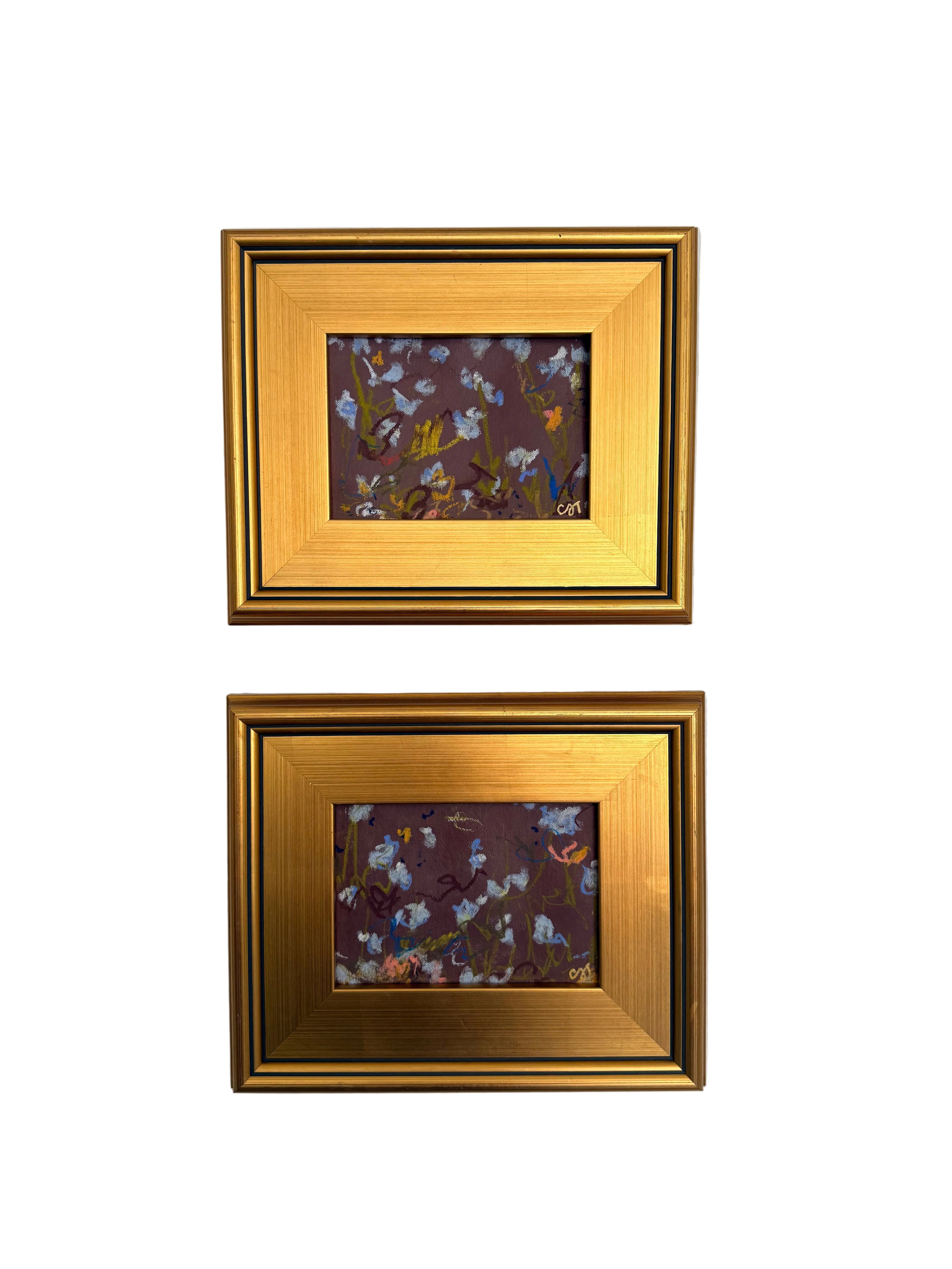 Two small framed artworks hanging vertically on a textured white wall. Each frame has a wide gold inner border and a narrow dark outer frame, enclosing a square floral painting with purple, blue, white, and yellow tones.
